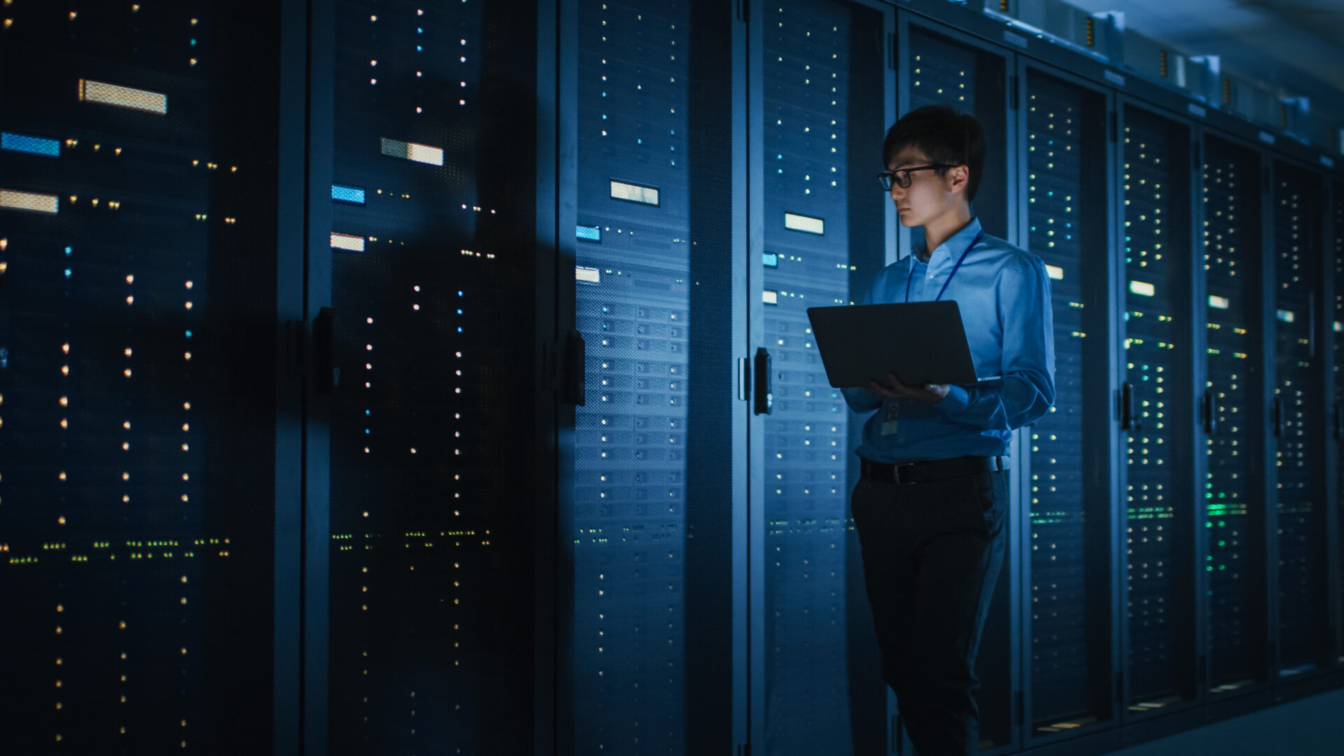 In Dark Data Center: Male IT Specialist Walks along the Row of Operational Server Racks, Uses Laptop for Maintenance. Concept for Cloud Computing, Artificial Intelligence, Supercomputer, Cybersecurity