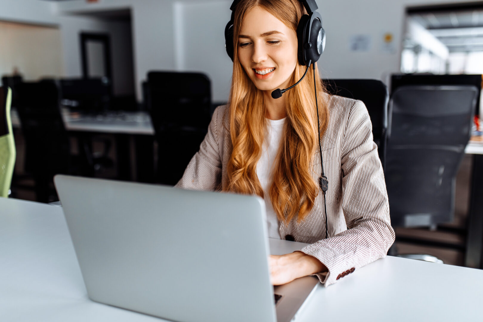 Smiling young female operator in headset works while sitting at the table, uses laptop, working in the call center.