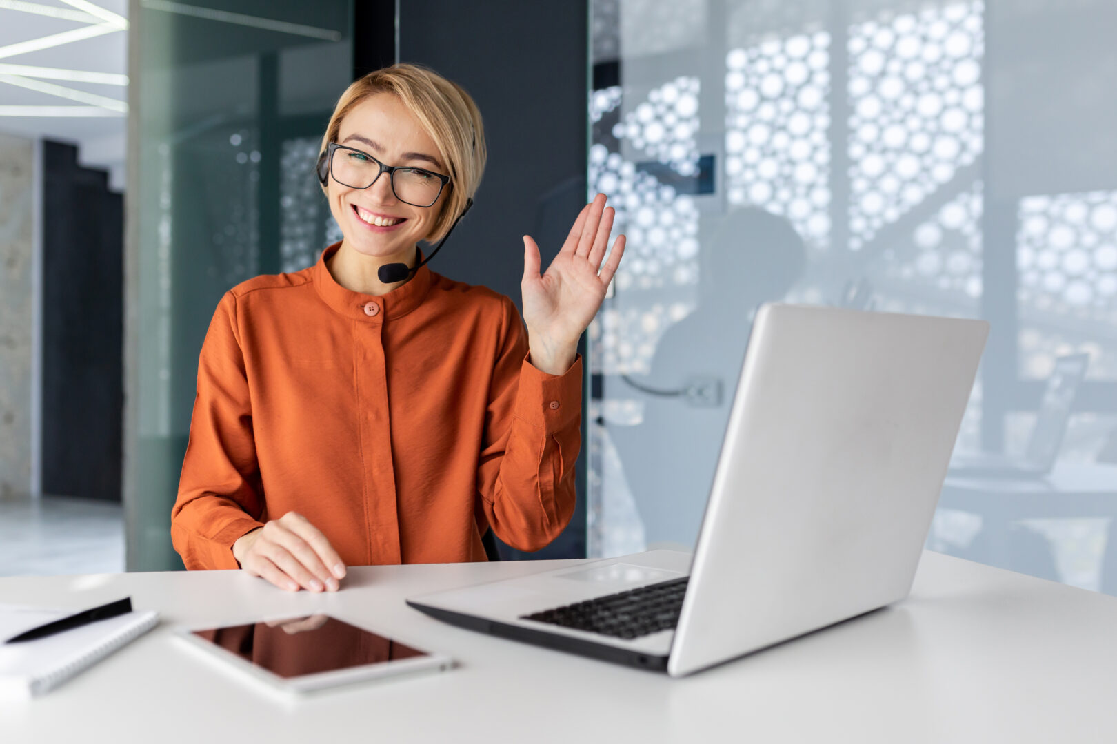 Portrait of successful tech support worker, woman with blonde hair smiling and looking at camera, businesswoman with headset for video call, greeting gesture, inside office with laptop